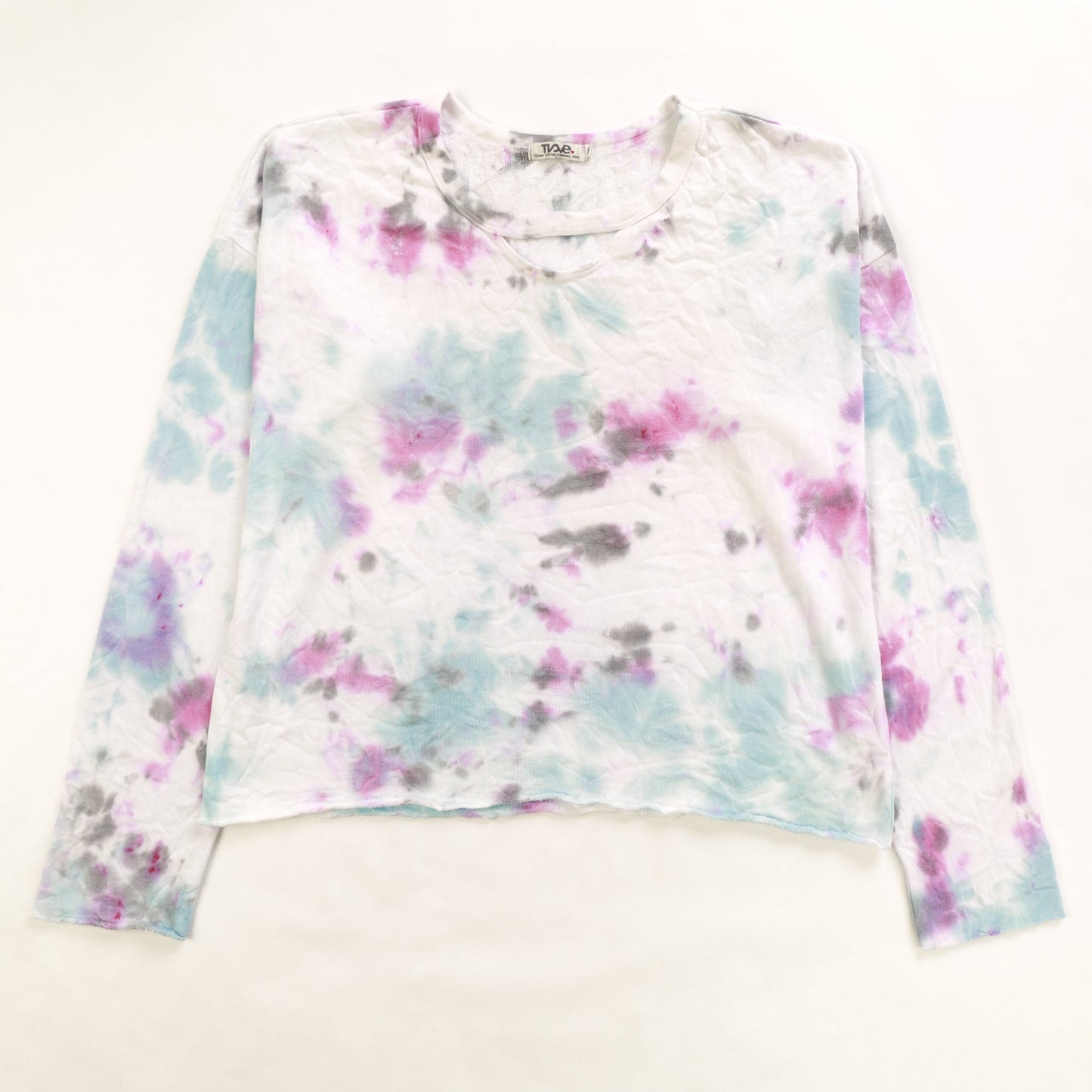 women's looser-fit long-sleeved shirt with raw edges at the sleeves and bottom hem and a half-circle keyhole cutout right underneath the neckline with blue, pink, and gray tie-dye splatters on white colored fabric